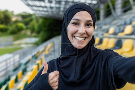 Téléchargez les photos : Femme souriante dans un hijab noir capture un selfie avec un pouce levé dans un stade de sport en plein air avec des sièges jaunes. - en image libre de droit