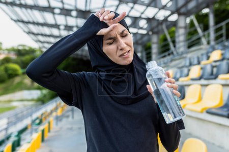 An exhausted female athlete in hijab wiping sweat from her forehead with a bottle of water at a stadium.