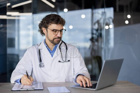 Téléchargez les photos : Un médecin professionnel en blouse de laboratoire utilisant un ordinateur portable et écrivant des notes sur un presse-papiers dans un cadre de soins de santé moderne. - en image libre de droit