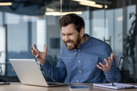An angry businessman is feeling frustrated with technology, showing stress and aggression towards his laptop in an office environment.