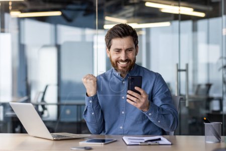Téléchargez les photos : Entrepreneur masculin triomphant se réjouit d'un succès commercial à son poste de travail moderne avec smartphone et ordinateur portable. - en image libre de droit