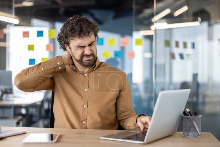 Un empleado de oficina masculino siente un dolor agudo en el cuello mientras trabaja en su computadora portátil, lo que indica la necesidad de espacios de trabajo ergonómicos.