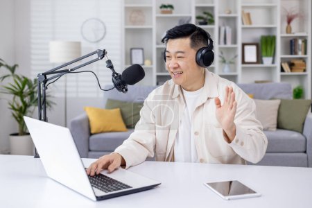 An engaging Asian man waving while recording a podcast using a laptop and microphone set up at his home office.