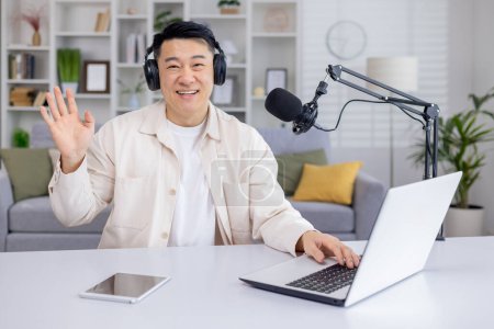 A cheerful male podcaster is greeting viewers with a wave while recording a live podcast session at his home studio setup.