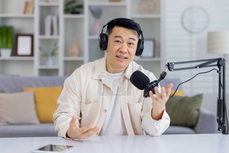 Friendly Asian man with headphones podcasting, talking and gesturing towards the camera with a broadcast microphone and a laptop in a home studio setting.