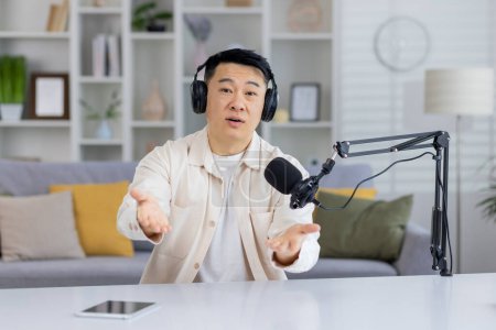 Photo for An engaging Asian man talking and looking at the camera while recording a podcast in a home studio setting. - Royalty Free Image