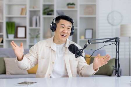 Enthusiastic Asian podcaster talking with hands, having an engaging conversation indoors, looking at camera with headphones and microphone.