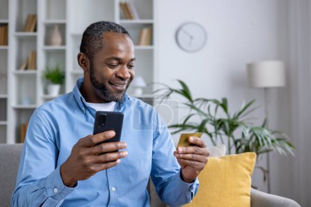 Téléchargez les photos : Homme décontracté utilisant smartphone et carte de crédit pour les achats en ligne tout en étant assis confortablement à la maison avec des plantes d'intérieur et intérieur lumineux. - en image libre de droit