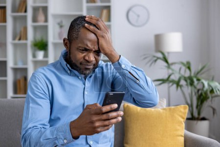 Anxious African American man feeling confused and worried while reading a message about fraud on his smartphone at home.