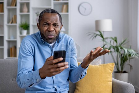 Middle-aged man sitting on couch, expressing confusion and disbelief while staring at his phone, looking at camera.
