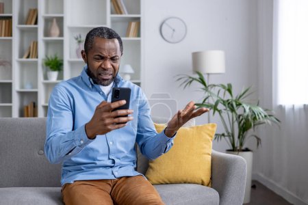 Ratloser afroamerikanischer Mann sitzt in einem Wohnzimmer, hält ein Smartphone in der Hand und gestikuliert verwirrt über eine trügerische Botschaft.
