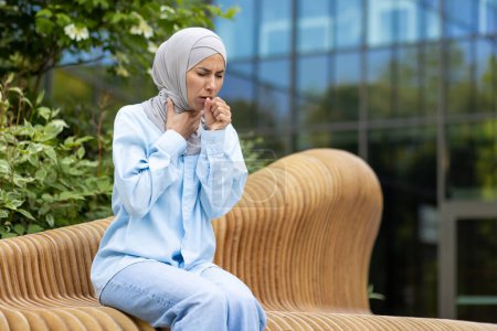 Muslim woman coughing and feeling sick while having pulmonary disease and sitting outdoors. Struggling lady covering mouth with clenched fist for not spreading virus around in downtown district.