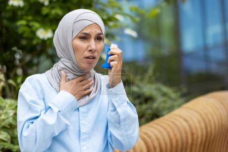 Téléchargez les photos : Femme arabe choquée couvrant le cou avec la main et tenant un inhalateur d'asthme bronchique tout en marchant à l'extérieur. Dame effrayée souffrant d'allergie saisonnière et se sentant difficile à respirer sans médicament. - en image libre de droit