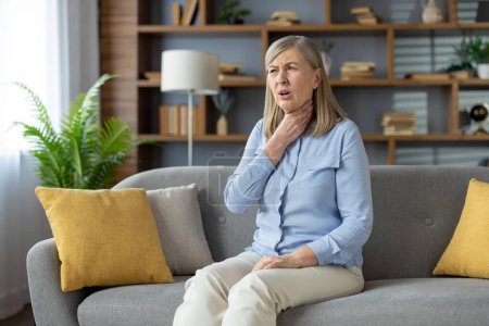 Sick woman sitting on couch with yellow pillows and suffering from sore throat while holding hand on neck. Unhealthy female having symptoms of flu or laryngitis disease while spending day at at home.