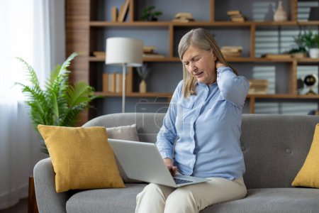 Müde ältere Dame im blauen Hemd reibt sich angespannten Nacken, während sie mit Laptop an den Beinen auf dem Sofa sitzt. Überlastetes weibliches Berühren des Hinterkopfes mit geschlossenen Wows zur Entlastung der Rückenmuskulatur.