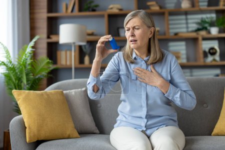 Grey haired woman in light outfit holding arm on chest while using medication for respiratory problem in living room. Disturbed patient experiencing asthma attack and relieving breathing struggles.