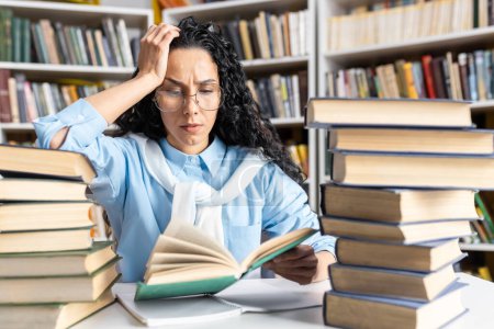 Overwhelmed Hispanic female student studying hard in a library. Piles of books surround her as she reads intently.