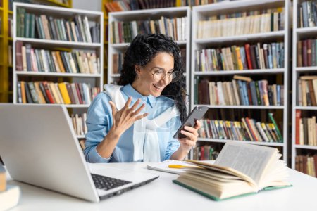 Foto de Alegre estudiante hispana con gafas que expresan felicidad mientras lee su smartphone, rodeada de libros en la biblioteca. - Imagen libre de derechos