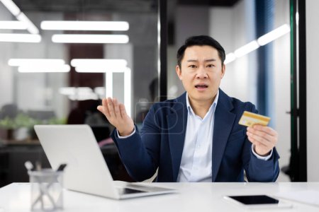 Photo for Asian businessman in office looks confused while holding a credit card and gesturing at camera, possibly dealing with fraud. - Royalty Free Image