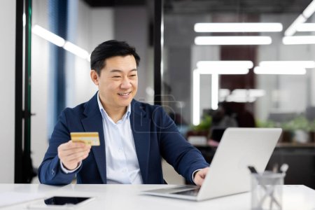 Professioneller und glücklicher asiatischer Geschäftsmann kauft online mit Kreditkarte und Laptop an seinem modernen Büroarbeitsplatz ein.