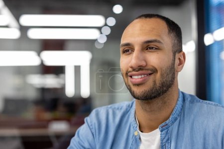 Ein professioneller hispanischer Mann mit freundlichem Auftreten, der in einem hellen, modernen Büroumfeld fotografiert wurde und Zuversicht und Positivität vermittelt.