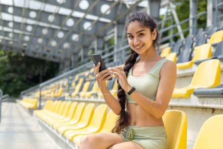 Foto de Retrato de una joven india sentada en ropa deportiva en el estadio tribuna, sosteniendo un teléfono móvil y sonriendo a la cámara. - Imagen libre de derechos