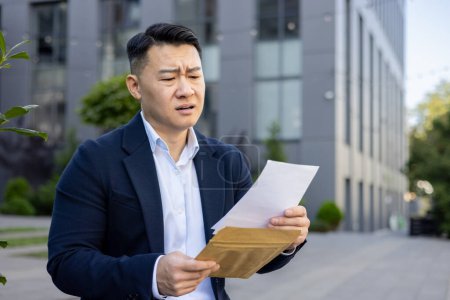 Upset and disappointed Asian businessman sitting outside the office and reading a received letter with bad news.