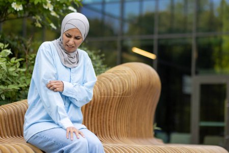 Foto de Mujer musulmana en hiyab sentada afuera, arañándose el brazo con una expresión dolorosa debido a una aparente reacción alérgica. - Imagen libre de derechos