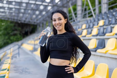 A fit Hispanic woman pausing to drink water during her exercise routine in an empty stadium.