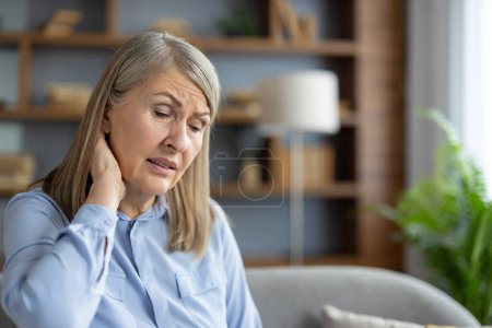 A senior Caucasian woman sitting in a living room appears distressed and uncomfortable, possibly experiencing neck pain or general malaise.