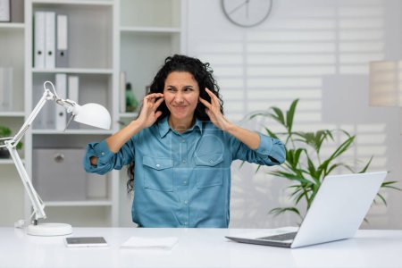 Una mujer profesional con una camisa azul sintiéndose abrumada por el ruido en su escritorio de la oficina con un ordenador portátil, plantas y una lámpara