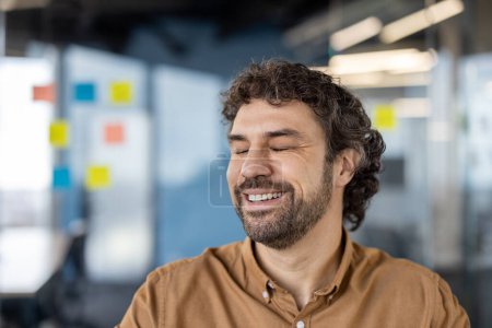 Foto de Un hombre alegre de mediana edad con el pelo rizado sonriendo ligeramente en un ambiente de trabajo casual. Parece contento y tranquilo, lo que sugiere un ambiente de oficina cómodo y positivo.. - Imagen libre de derechos