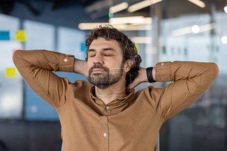 Ein Mann mittleren Alters mit lockigem Haar entspannt sich, indem er die Arme hinter dem Kopf in einer modernen Büroatmosphäre ausbreitet und einen Moment der Stressabbau demonstriert.