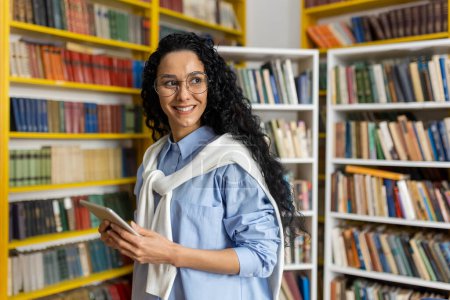 Photo for A cheerful curly-haired woman stands with a tablet in a vibrant library. She is wearing glasses and a casual blue outfit, exploring academic resources. - Royalty Free Image