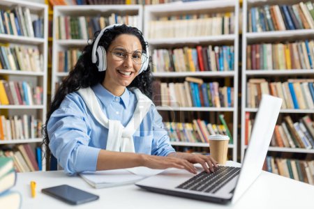 Foto de Mujer sonriente en un entorno de biblioteca usando un portátil con auriculares. Está rodeada de amplias estanterías, ejemplificando un entorno productivo de investigación.. - Imagen libre de derechos