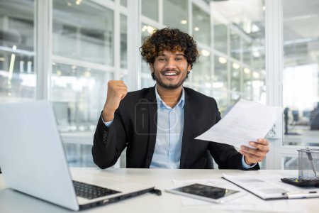 Photo for A cheerful young man in a business suit celebrates a successful project or achievement while sitting at his desk in a modern office environment, showing victory fist and smiling with documents in hand - Royalty Free Image