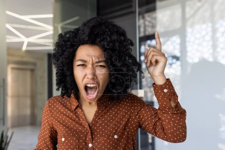 African American woman pointing upwards and shouting, portraying frustration or an urgent explanation during a video call in a modern office.