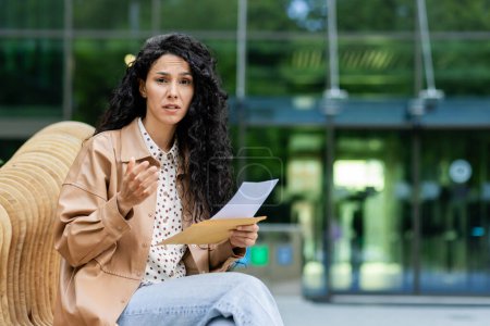 A young Hispanic woman sits outdoors, reading a document with a concerned expression. Set against a modern cityscape backdrop, she appears engaged and somewhat worried.