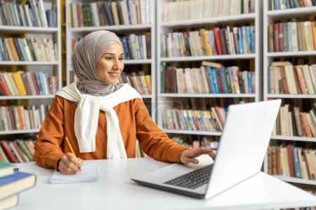A cheerful Muslim woman wearing hijab works diligently on a laptop in a library setting, surrounded by an array of books.