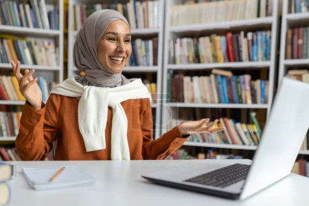 A vivacious young woman wearing a hijab is smiling while engaging in an animated video call. She sits in a library, surrounded by books, exuding a friendly demeanor.
