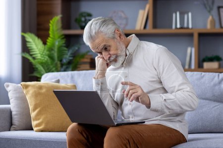 Focused elderly man with white hair and beard deeply engrossed in using a laptop while sitting on a sofa in a well-lit living room, depicting modern technology use by seniors.