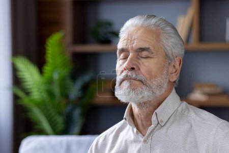 Portrait serein d'un aîné aux cheveux gris méditant les yeux fermés, assis confortablement dans son salon bien décoré avec des plantes vertes vibrantes.