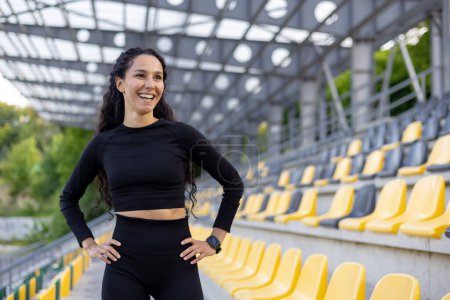 Foto de Mujer hispana alegre en ropa deportiva descansando y sonriendo en un estadio deportivo. Fitness, alegría y un concepto de estilo de vida activo capturados bellamente al aire libre. - Imagen libre de derechos