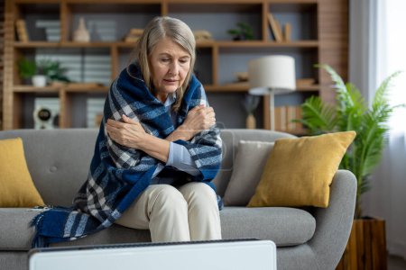 Kranke Seniorin umarmt sich, während sie mit einer karierten Decke bedeckt neben dem elektronischen Heizkörper sitzt. Besorgtes Weibchen versucht warm zu werden, während es auf den Beginn der Heizsaison wartet.