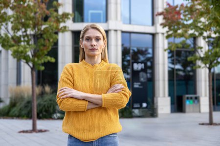 Eine nachdenkliche Frau mit verschränkten Armen in einem leuchtend gelben Pullover steht selbstbewusst in einem urbanen Outdoor-Umfeld und vermittelt Professionalität und Entschlossenheit.