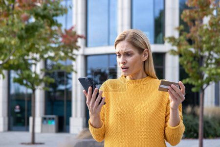 Puzzled blonde woman in yellow sweater holds credit card and phone while standing outside a modern glass building, appearing concerned and confused.