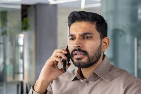 Téléchargez les photos : Un Indien a l'air tendu et frustré en parlant sur son téléphone portable dans un bureau moderne, montrant des signes de colère et de stress. - en image libre de droit