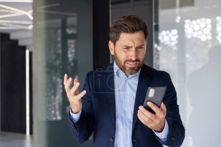 Téléchargez les photos : Homme d'affaires d'âge moyen exprimant sa frustration en regardant son smartphone dans un environnement de bureau élégant et moderne, rayonnant de stress et d'urgence. - en image libre de droit