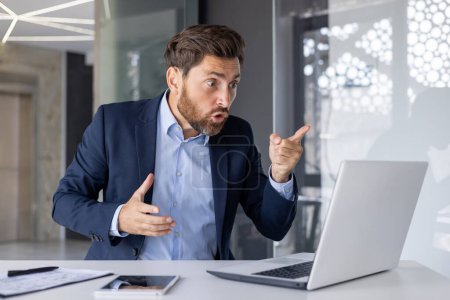 Frustrated male executive in a business suit engaged in a heated discussion over a video call, pointing and gesturing angrily in a modern office setting.