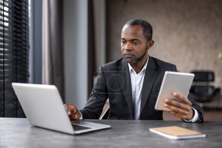 Konzentrierter afroamerikanischer Geschäftsmann nutzt Laptop und Tablet an seinem modernen Arbeitsplatz, um Engagement und Technologieeinsatz in einem professionellen Umfeld darzustellen.